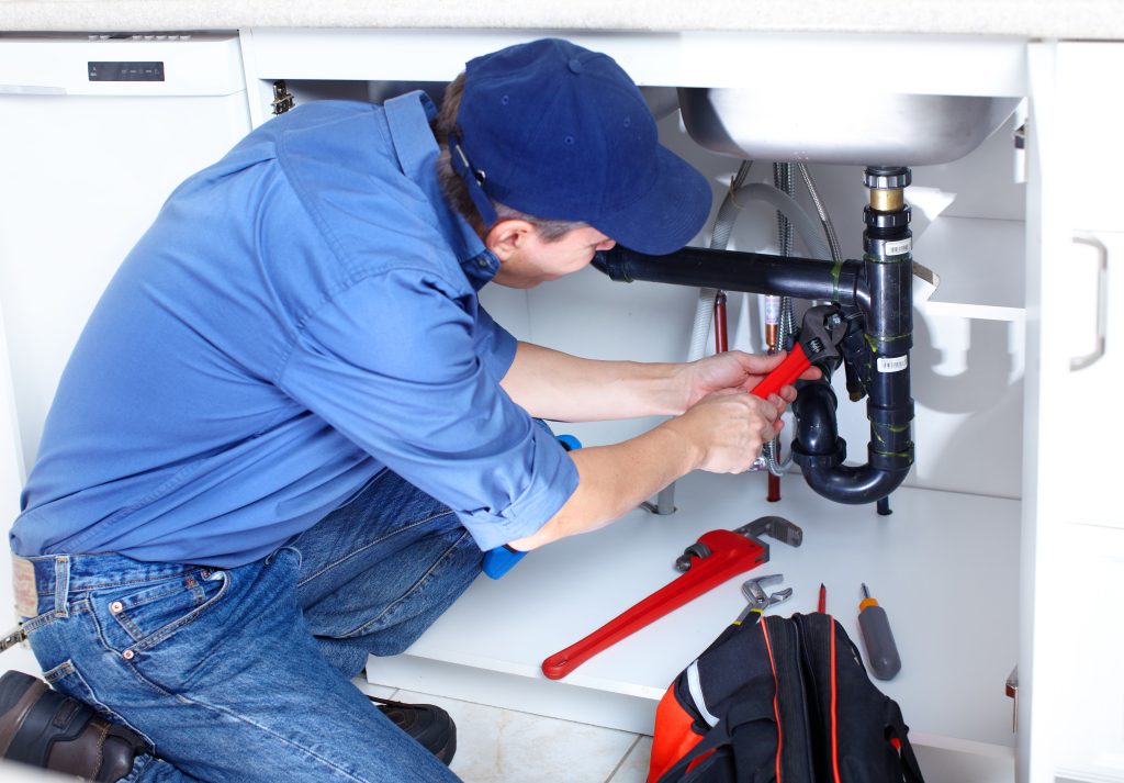 A plumber in a blue uniform and cap working under a kitchen sink, using a wrench to fix the plumbing, with various tools laid out on the floor nearby.