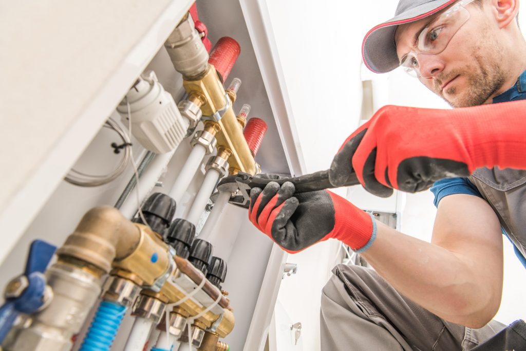 A technician wearing red gloves and safety glasses uses a wrench to tighten a connection on a set of metal pipes connected to a plumbing system.