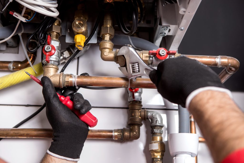 Close-up of a technician's hands wearing black gloves while using a wrench and a screwdriver to install or repair a heating system. The image shows various pipes, valves, and connections, including red and yellow shut-off valves