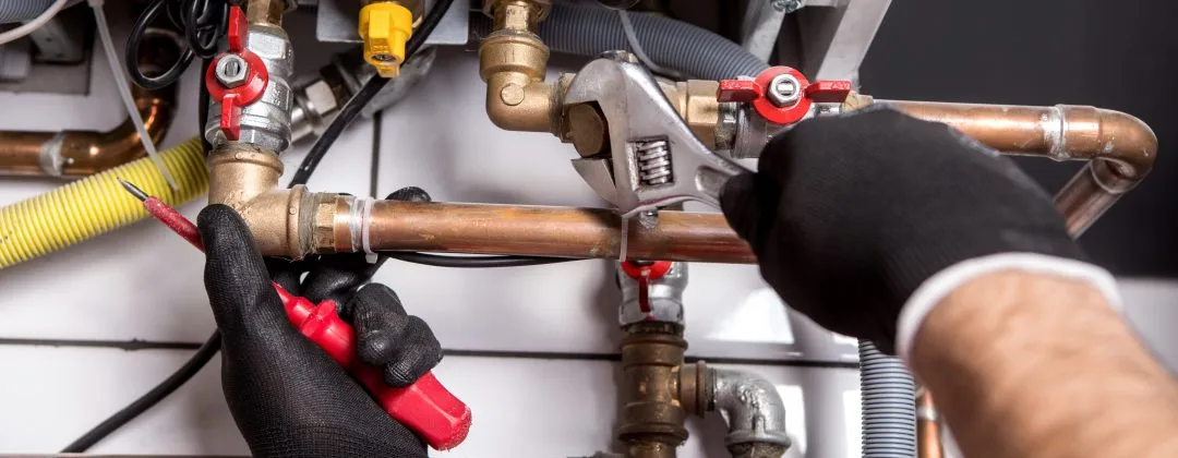 Close-up of a technician's hands wearing black gloves while using a wrench and a screwdriver to install or repair a heating system. The image shows various pipes, valves, and connections, including red and yellow shut-off valves